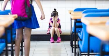 Menina em pé na sala de aula fazendo bullying com sua colega de classe, que chora no chão, sentada.