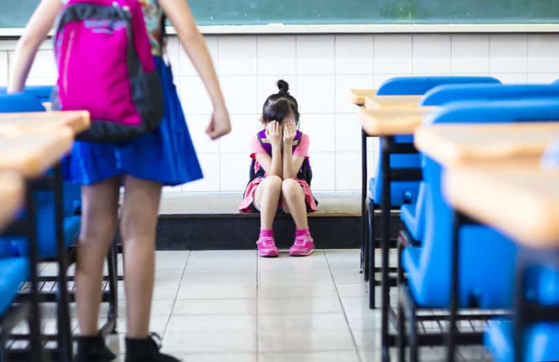 Menina em pé na sala de aula fazendo bullying com sua colega de classe, que chora no chão, sentada.