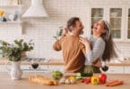 Casal dançando alegremente na cozinha, com alimentos dispostos na bancada à sua frente.