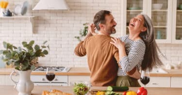 Casal dançando alegremente na cozinha, com alimentos dispostos na bancada à sua frente.