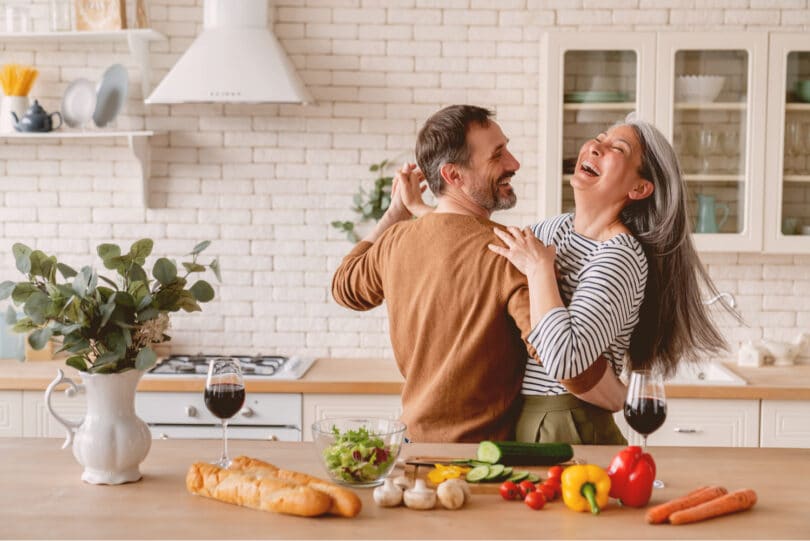 Casal dançando alegremente na cozinha, com alimentos dispostos na bancada à sua frente.