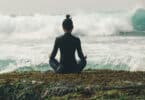 Mulher sentada em pose de meditação, de costas, mas de frente para o mar com ondas fortes.