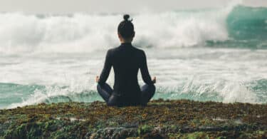 Mulher sentada em pose de meditação, de costas, mas de frente para o mar com ondas fortes.