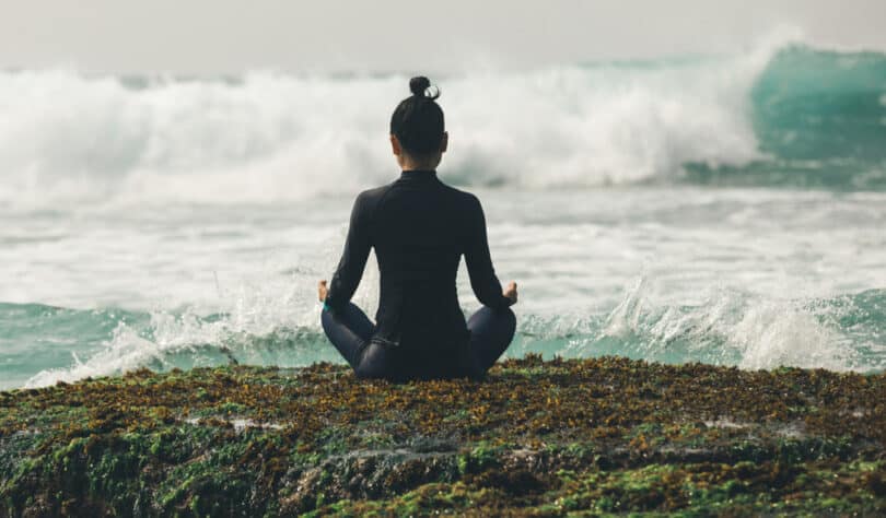 Mulher sentada em pose de meditação, de costas, mas de frente para o mar com ondas fortes.