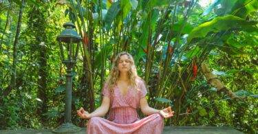 Mulher loira meditando sentada, com as mãos sobre seus joelhos. Ao seu redor, uma paisagem verde, cheia de árvores.