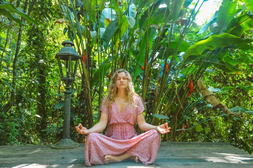 Mulher loira meditando sentada, com as mãos sobre seus joelhos. Ao seu redor, uma paisagem verde, cheia de árvores.