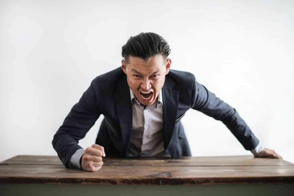 Homem com roupas sociais gritando enquanto bate com o punho na mesa de escritório.