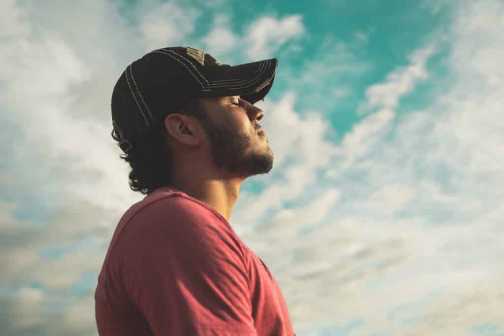 Homem de boné e camiseta vermelha de olhos fechados ao ar livre. 