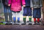 Foto de quatro crianças, três meninos e uma menina, só da cintura para baixo, todos vestindo roupas de frio coloridas e galochas de chuva.