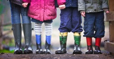 Foto de quatro crianças, três meninos e uma menina, só da cintura para baixo, todos vestindo roupas de frio coloridas e galochas de chuva.
