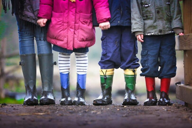 Foto de quatro crianças, três meninos e uma menina, só da cintura para baixo, todos vestindo roupas de frio coloridas e galochas de chuva.