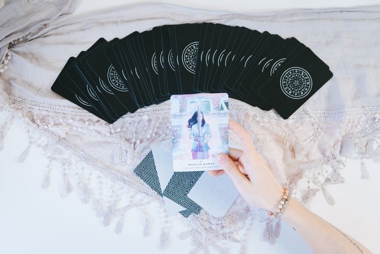 Baralho de tarot com todas as cartas pretas colocado em cima de uma mesa de mármore branco. Em frente é possível ver o braço de uma pessoa branca segurando uma carta, mostrando seu desenho em cores azuis.