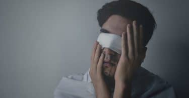 Homem jovem, vestindo camiseta branca, usando uma venda branca nos olhos, com as duas mãos tocando o rosto.