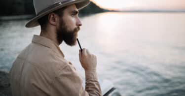 Homem sentado à beira de um rio, com uma caneta no queixo e pensando. Em seu colo, há um caderno baerto com folhas em branco