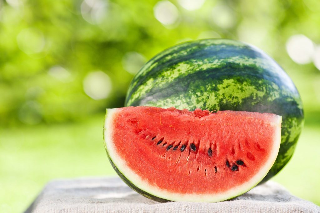 Melancia inteira sobre uma mesa e ao lado uma fatia da fruta já pronta para ser consumida. Ao fundo uma folhagem verde.
