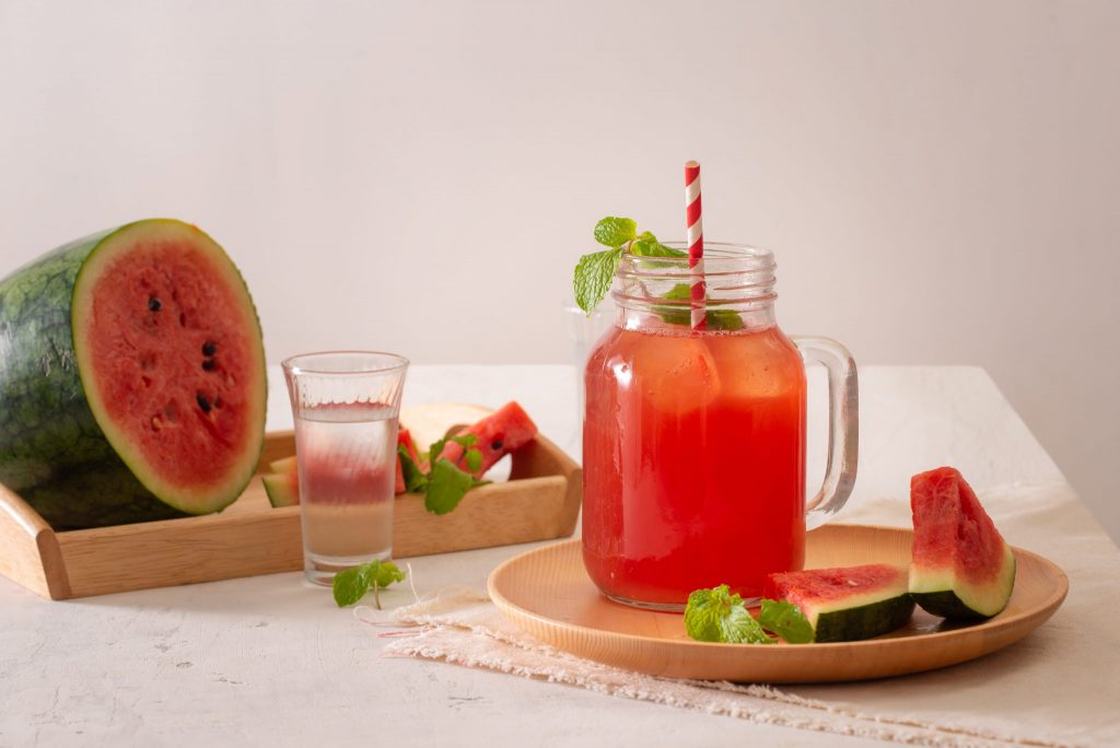 Jarra grande com suco detox de melancia. A jarra está sobre uma mesa e ao lado temos uma bandeja com metade de uma melancia e um poco de água. Também compõe a imagem, folhas de hortelã.