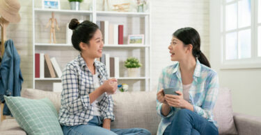 Duas mulheres sentadas no sofá conversando enquanto seguram xícaras com bebida.