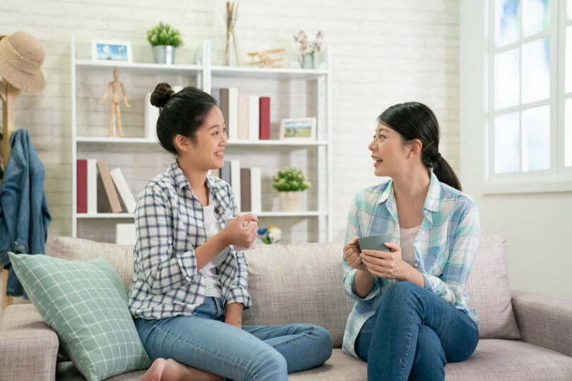 Duas mulheres sentadas no sofá conversando enquanto seguram xícaras com bebida.