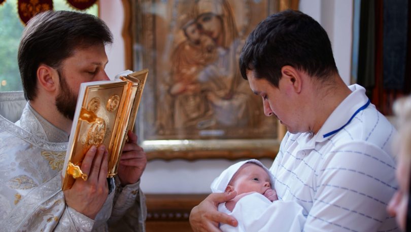 Homem segura bebê durante batizado, enquanto padre beija a bíblia.