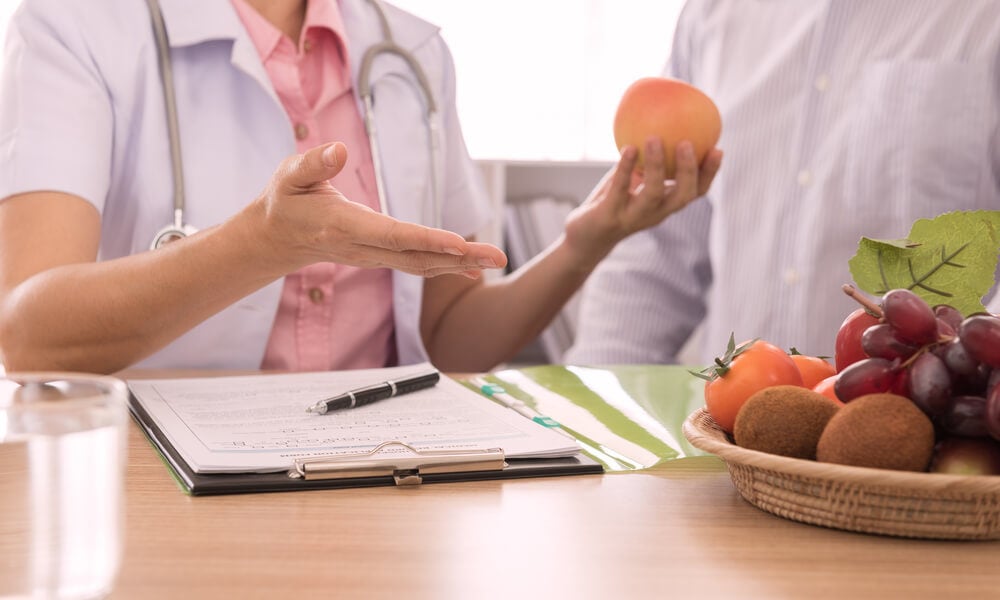 Imagem de uma médica com o seu paciente. Ela está expliando métodos de dieta. Ela segura uma fruta em suas mãos. Sobre a sua mesa encontra-se uma prancheta com anotações e uma fruteira com uvas, caquis e kiwis.