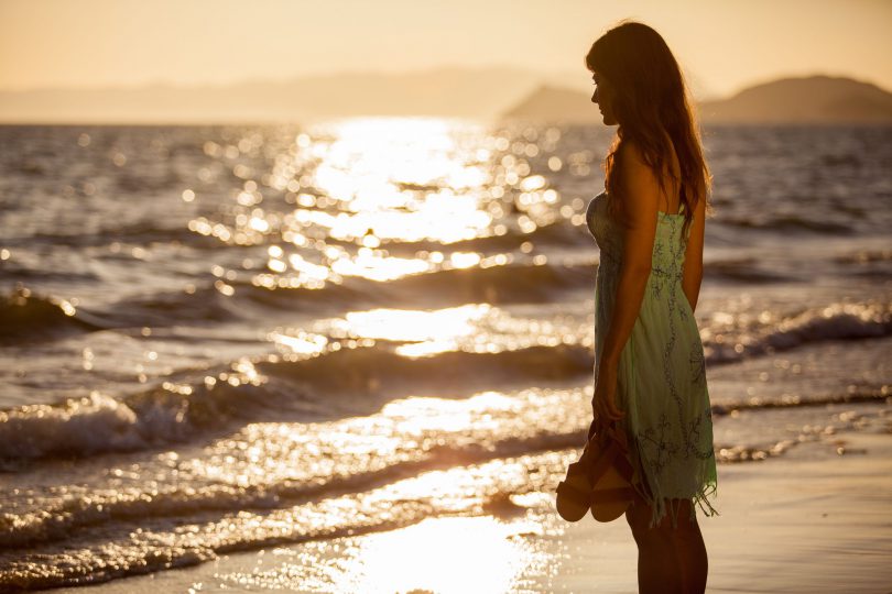 Mulher na beira da praia com chinelos na mão