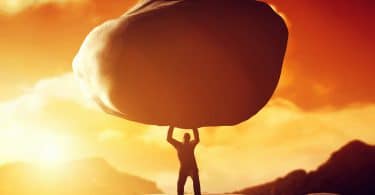 Silhueta de pessoa forte segurando uma pedra enorme sobre a sua cabeça. Ao fundo o céu laranja graças ao pôr do sol.