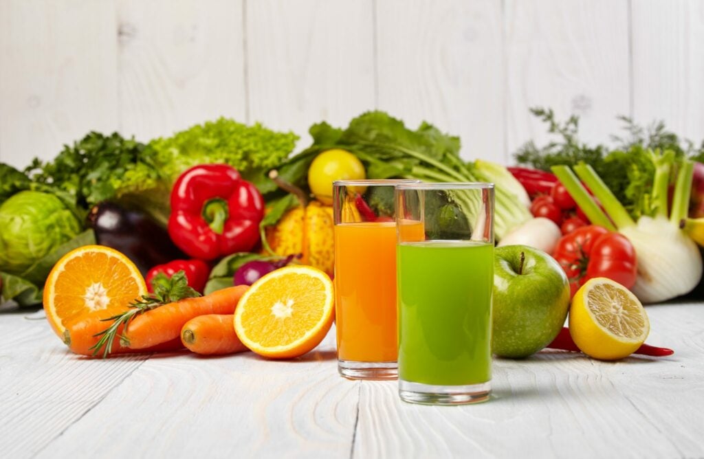 Diverso vegetais e frutas dispostos sobre uma mesa de tábua branca junto com dois copos cheios de suco, sendo um na cor amarela e outro na cor verde.
