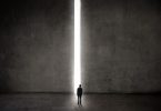Homem parado em frente a parede gigante de concreto, com abertura no meio por onde passa um feixe de luz.