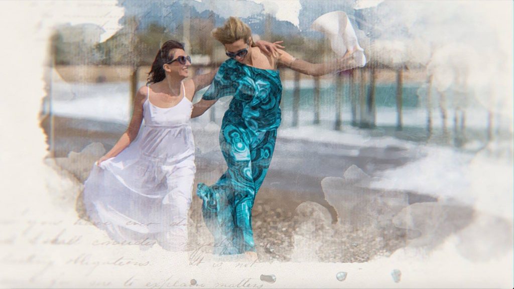 Duas mulheres caminhando sobre a areia do mar. Ambas estão vestindo um vestido longo, sendo um na cor branca e o outro na cor azul. Elas também usam óculos de sol.