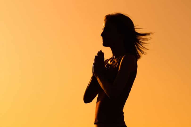 Silhueta de mulher sobre fundo laranja com as mãos sobre o peito em sinal de gratidão.