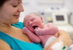Mulher branca e jovem em sala de parto segurando um bebê recém nascido.