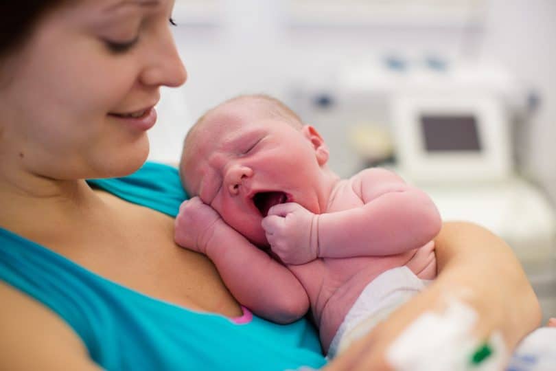 Mulher branca e jovem em sala de parto segurando um bebê recém nascido.