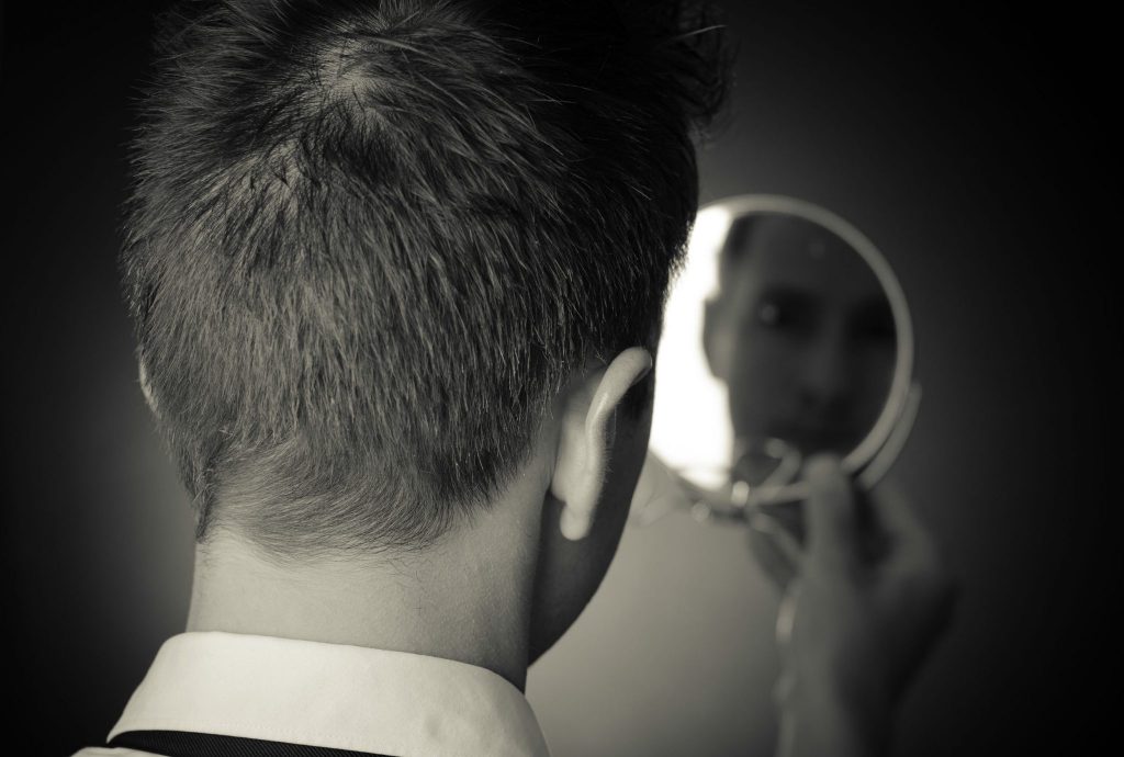 Foto preta e branca de homem de costas, vestindo social, observando o reflexo desfocado do seu rosto em um espelho pequeno e redondo