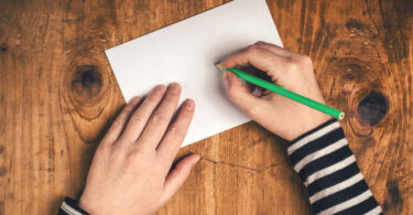 Mãos escrevendo em papel com lápis verde visto de cima