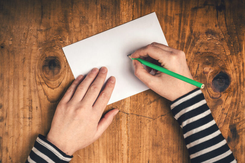 Mãos escrevendo em papel com lápis verde visto de cima