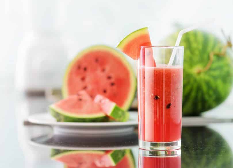 Suco de melancia em copo com canudo. Ao fundo, há melancia fatiada.