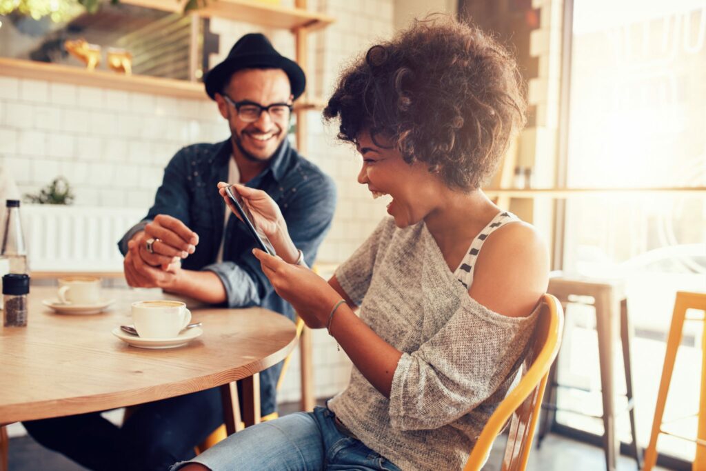 Mulher sorrindo em um café, olhando para um tablet. Ao seu lado, um amigo também ri.