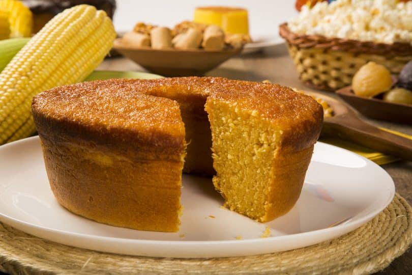 Bolo de milho com uma fatia cortada. Ao fundo, há espigas de milho e outros alimentos dentro de recipientes abertos.