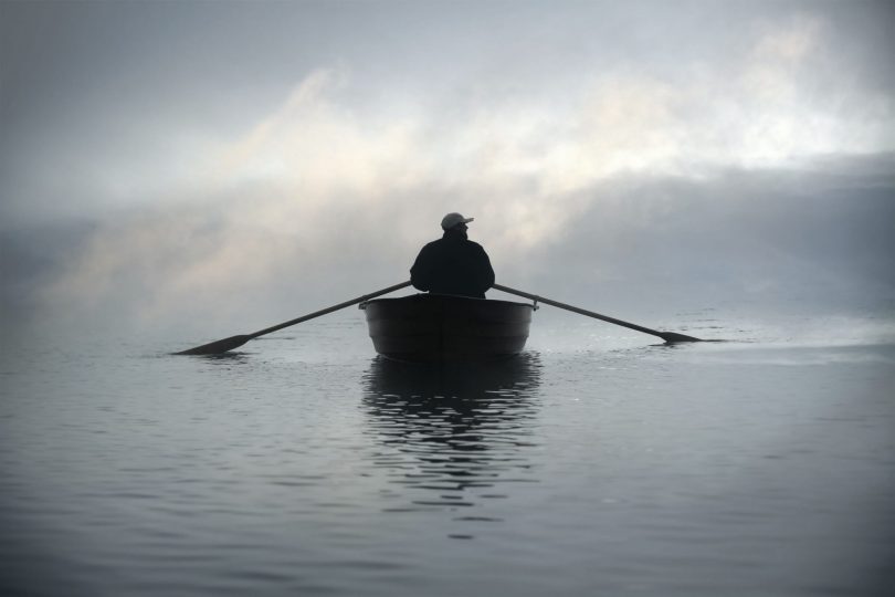 Foto de silhueta de pessoa em um barco, remando no meio do mar com o céu coberto por neblina.