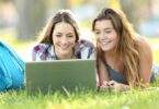 Duas meninas colegiais deitadas na grama, sorrindo e mexendo em notebook..