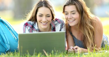 Duas meninas colegiais deitadas na grama, sorrindo e mexendo em notebook..