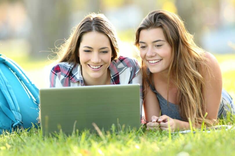 Duas meninas colegiais deitadas na grama, sorrindo e mexendo em notebook..