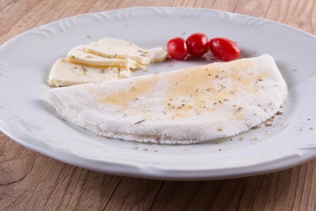 Tapioca pronta para ser servida disposta em um prato branco. Tomates cerejas e pedaços de queijo fresco decoram o prato. 