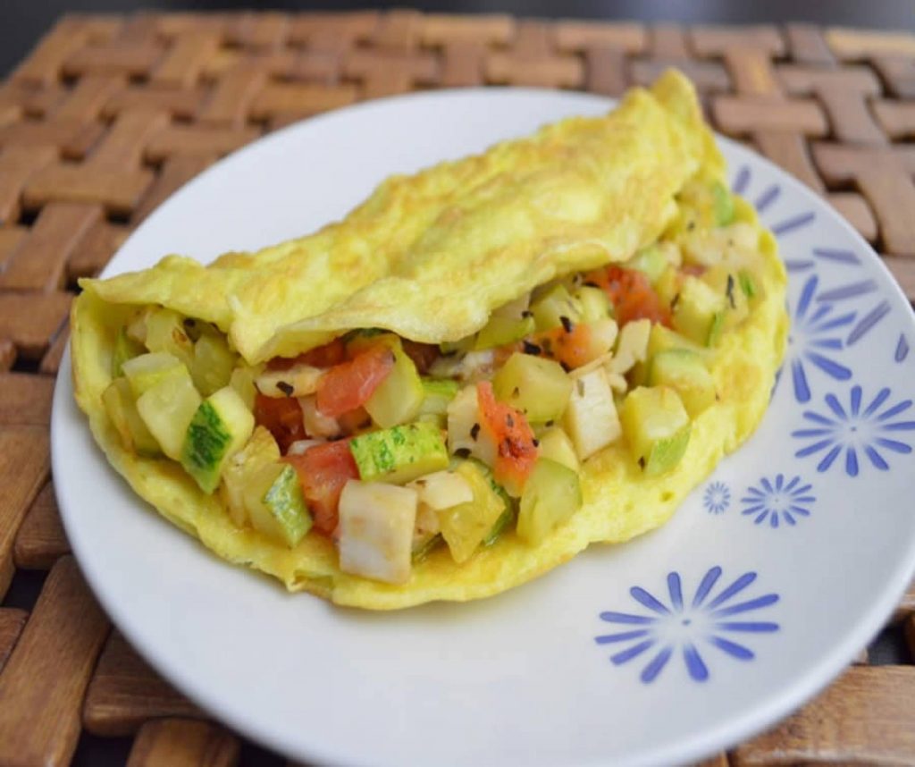 Tapioca recheada com abobrinhas picadinas e tomates cereja. Está disposta em um prato branco decorado com flores azul.