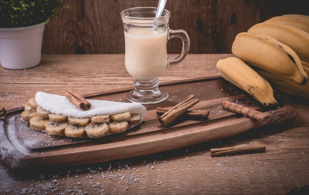 Tapioca recheada com bananas em rodelas. Está disposta em uma tábua de madeira decorada com canelas, bananas e um copo com suco.