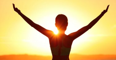 Silhueta de mulher com os braços abertos de frente para o o sol nascendo.