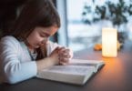 Menina pequena orando debruçada em cima de uma bíblia.