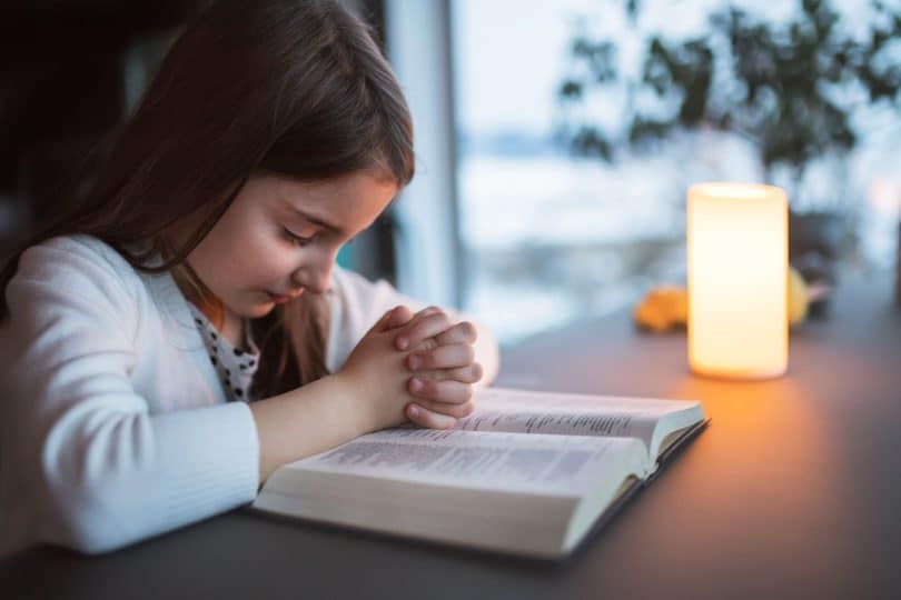 Menina pequena orando debruçada em cima de uma bíblia.
