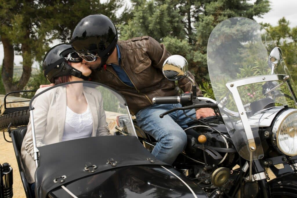 Casal de homem e mulher se beijando na boca. Ambos estão de capacete preto, óculos e sentados sobre uma moto.