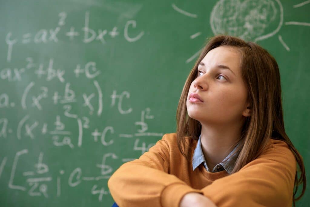 menina jovem, no ensino médio, sentada em frente de lousa.